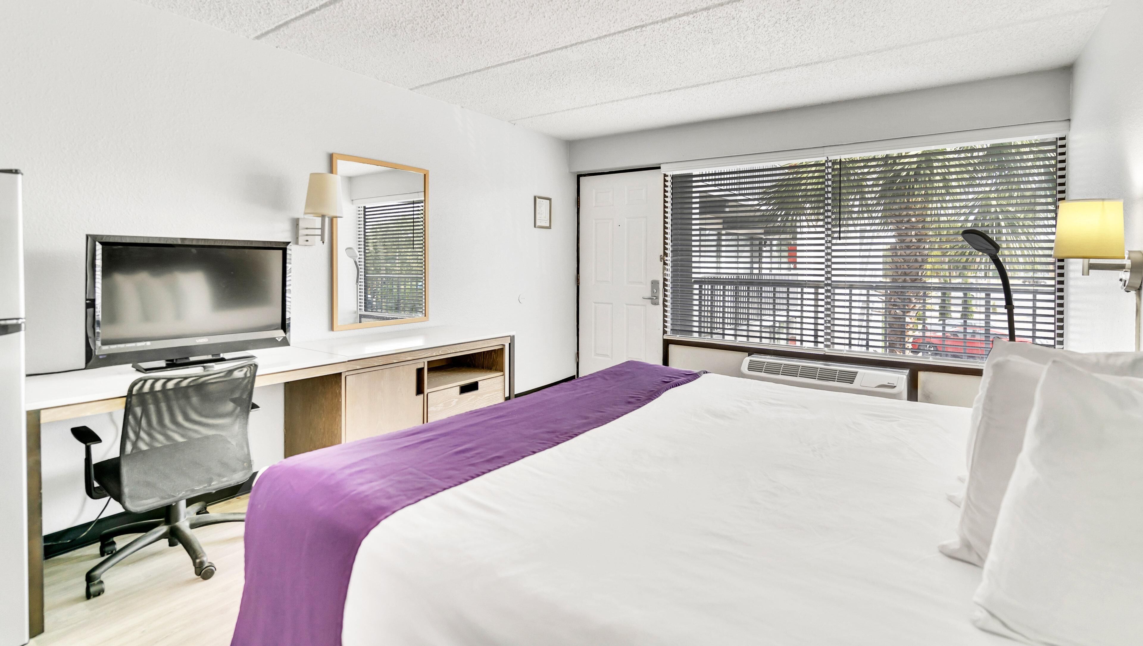 A hotel room with a bed, a desk with a computer, a chair, a TV, and a window with blinds.