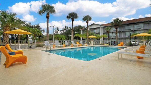 A swimming pool with lounge chairs and umbrellas, surrounded by palm trees and a building with balconies. - Home 1 Suites Extended Stay - Kissimmee (Kissimmee)