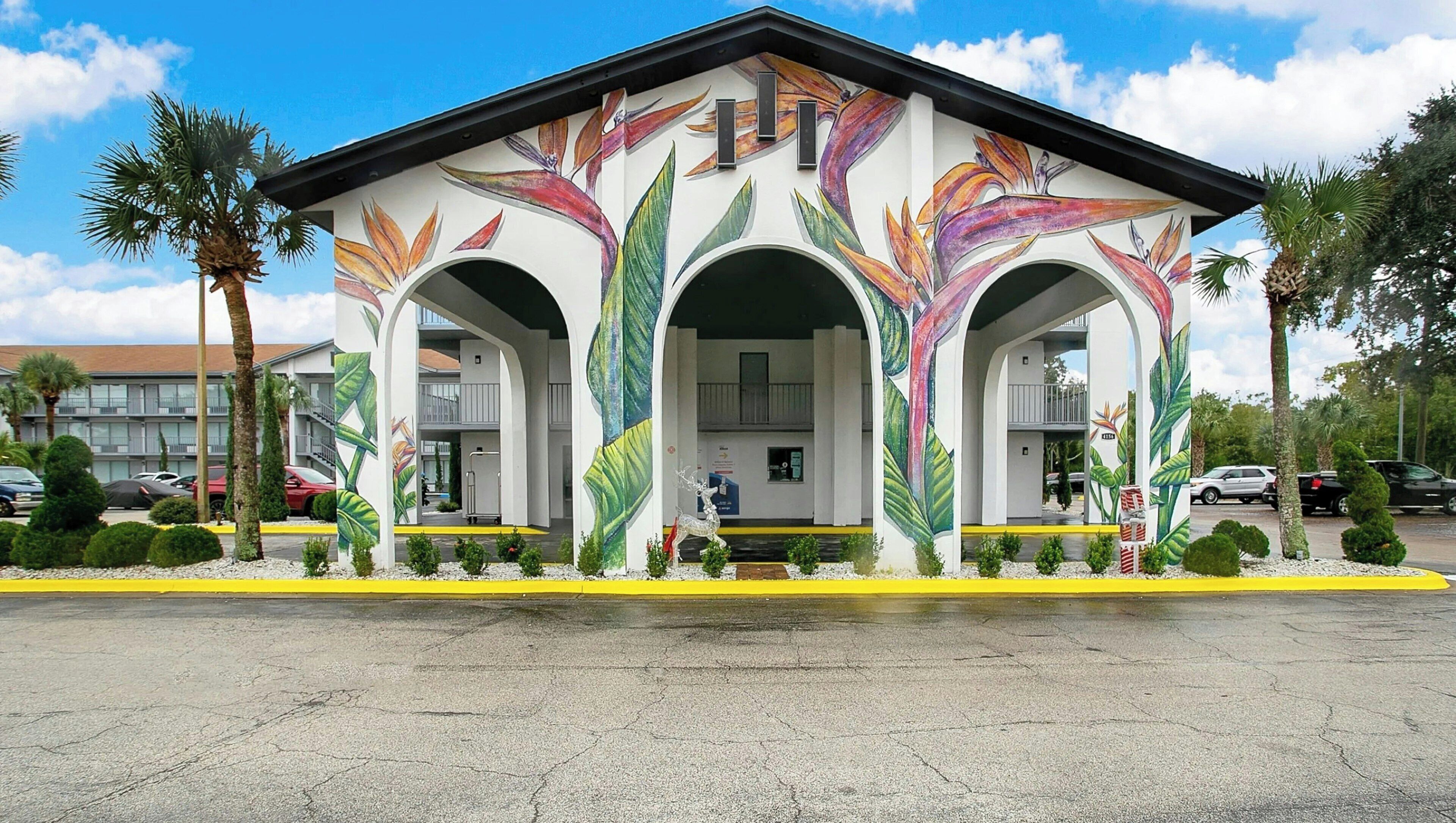 A building with a white facade and colorful tropical leaf murals, featuring three arches and a central entrance.