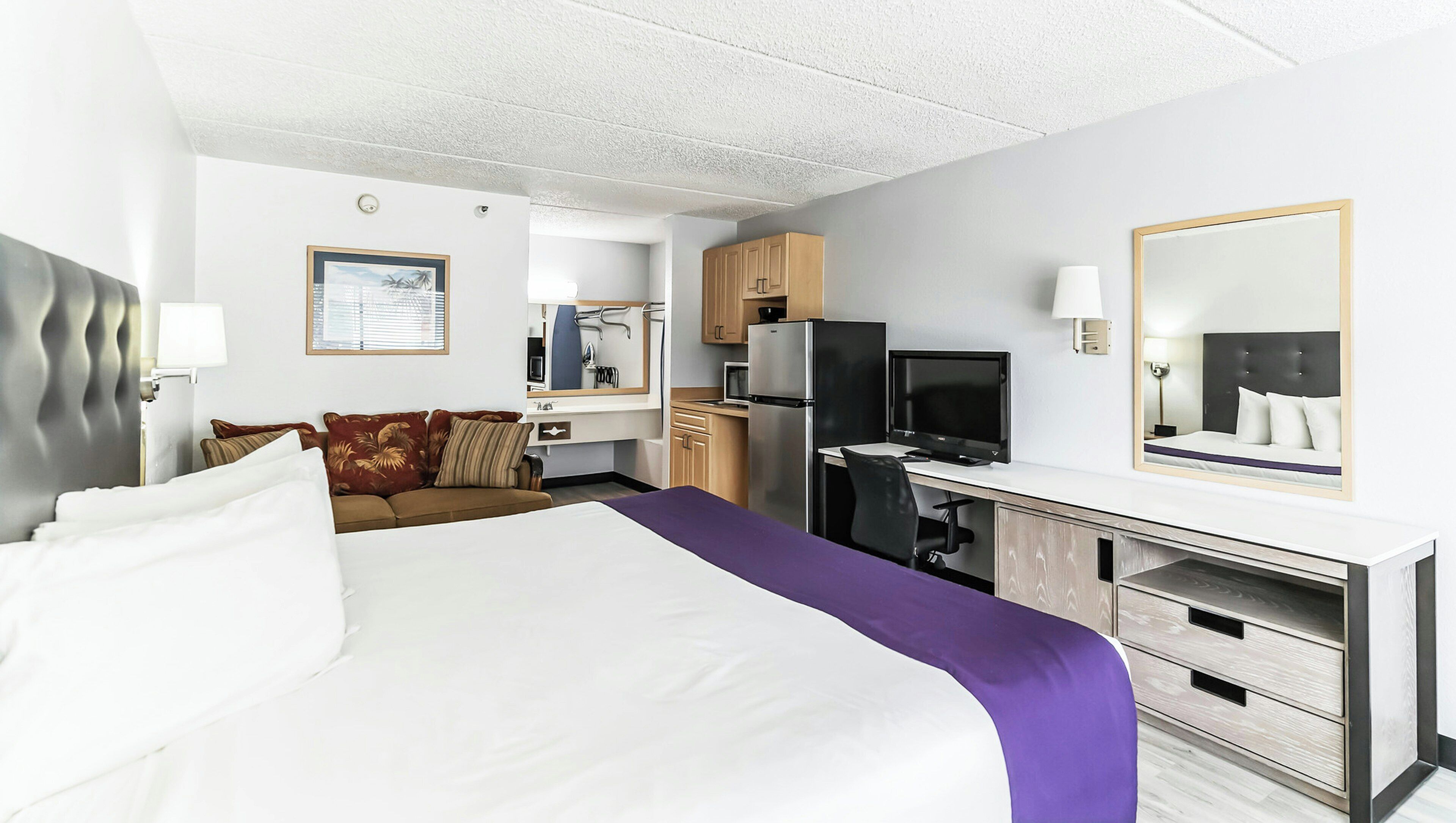 A hotel room with a bed, a desk, a television, a mirror, and a kitchenette.