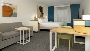 Una habitación de hotel con cama, sofá, mesita, sillón, televisor y un cuadro en la pared.