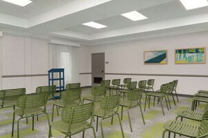 Un salón de conferencias con sillas verdes dispuestas en semicírculo, una mesa azul y dos cuadros abstractos en la pared.