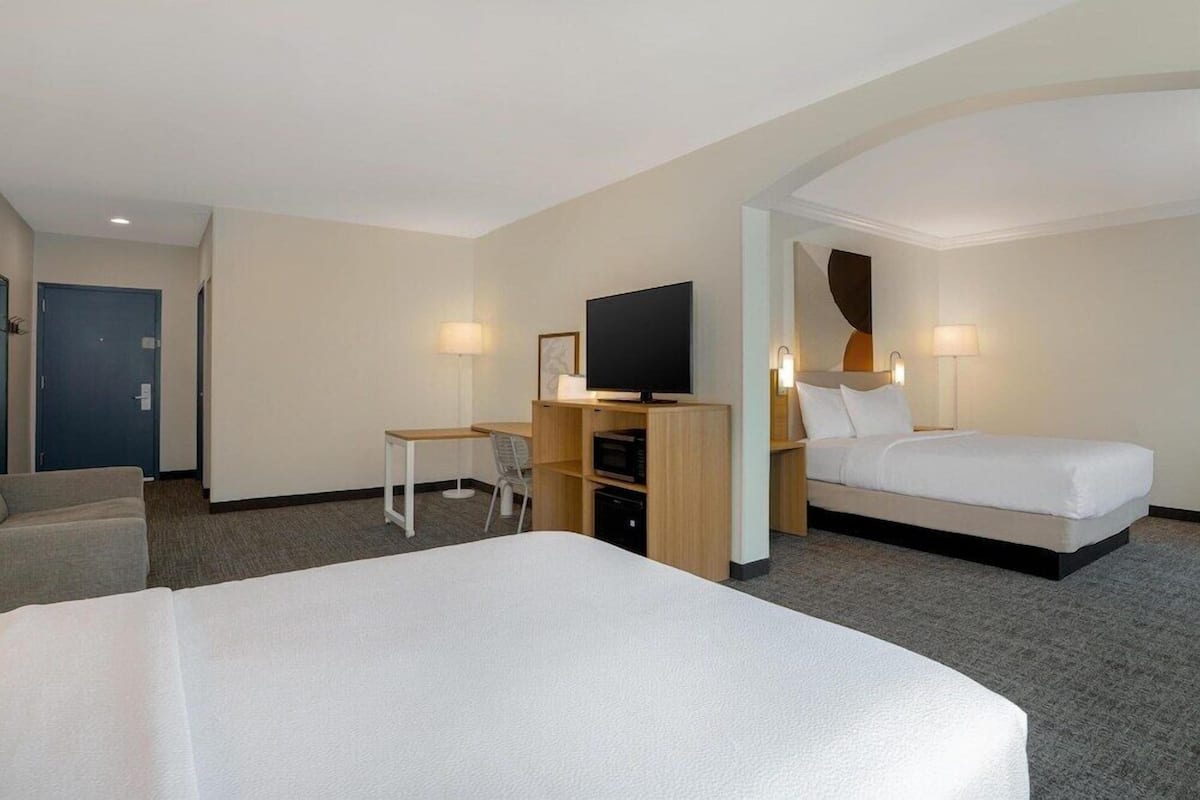 Habitación de hotel con una cama grande, una cama más pequeña, un escritorio con silla, un televisor y una puerta.