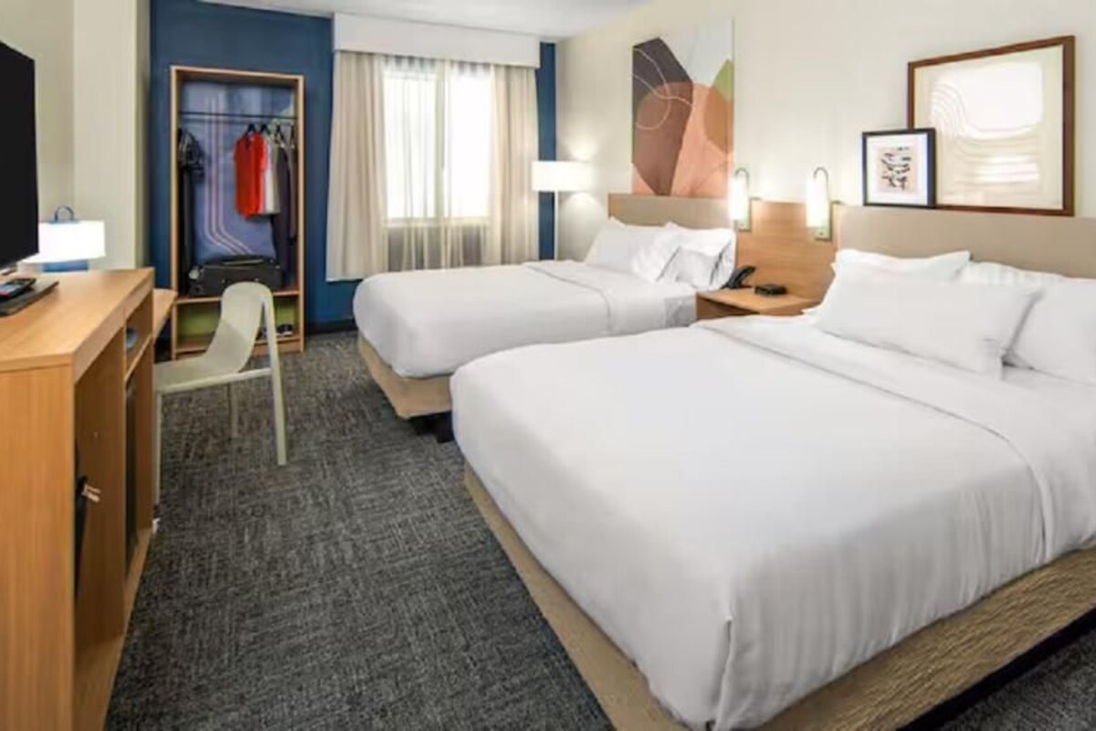 Une chambre d’hôtel avec deux lits, un bureau, une chaise, une armoire et une fenêtre avec des rideaux.