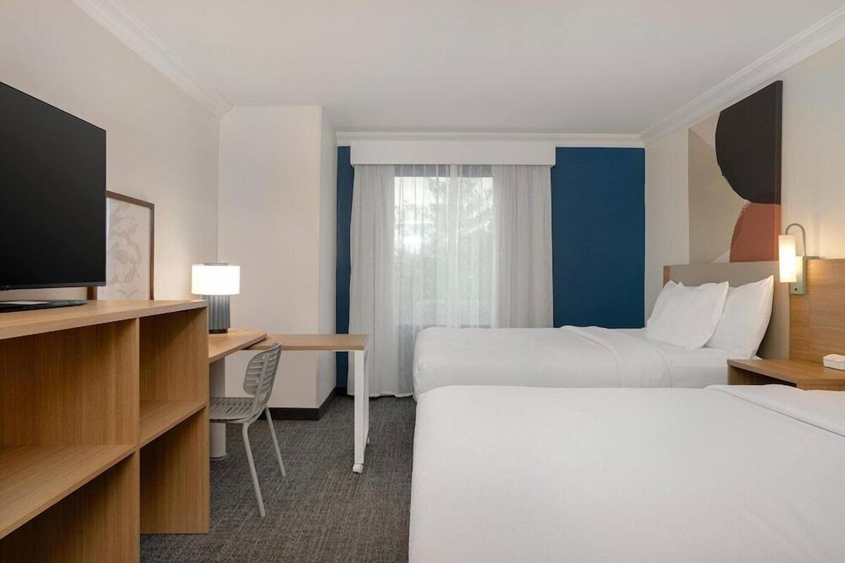 A hotel room with two beds, a desk, a chair, a TV, and a window with curtains.