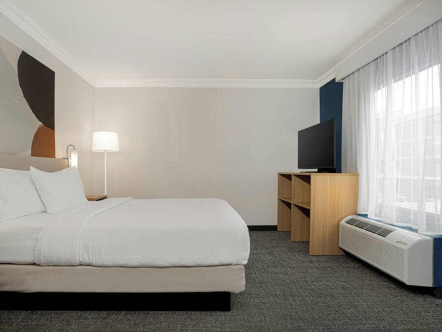 Habitación de hotel con cama, lámparas de noche, televisor y ventana con cortinas.