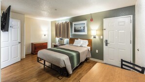 A bedroom with a bed, bedside table, lamp, and a door. - Hometowne Studios Atlanta - Chamblee (Chamblee)