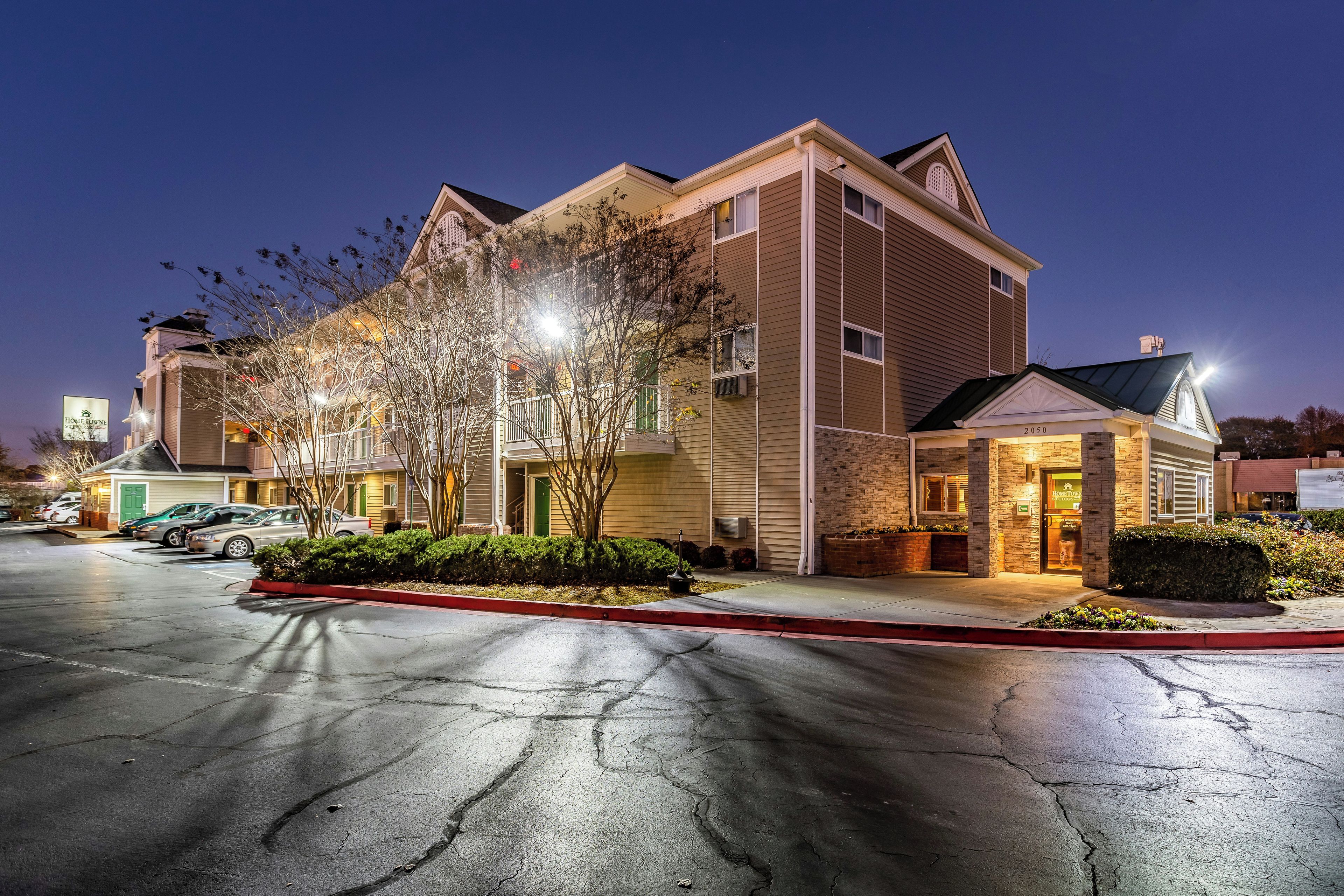 A multi-story hotel with a parking lot in front, featuring a well-lit entrance and a visible storefront.
