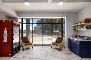 A room with a Coca-Cola refrigerator, two patterned armchairs, a large window, and a shelf with snacks. - Suburban Studios Hinesville - Fort Stewart Area (Hinesville)