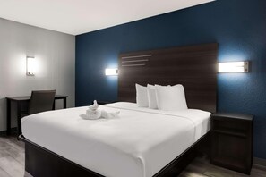 A hotel room with a large bed, bedside tables, a desk, and wall-mounted lights. - Suburban Studios Hinesville - Fort Stewart Area (Hinesville)