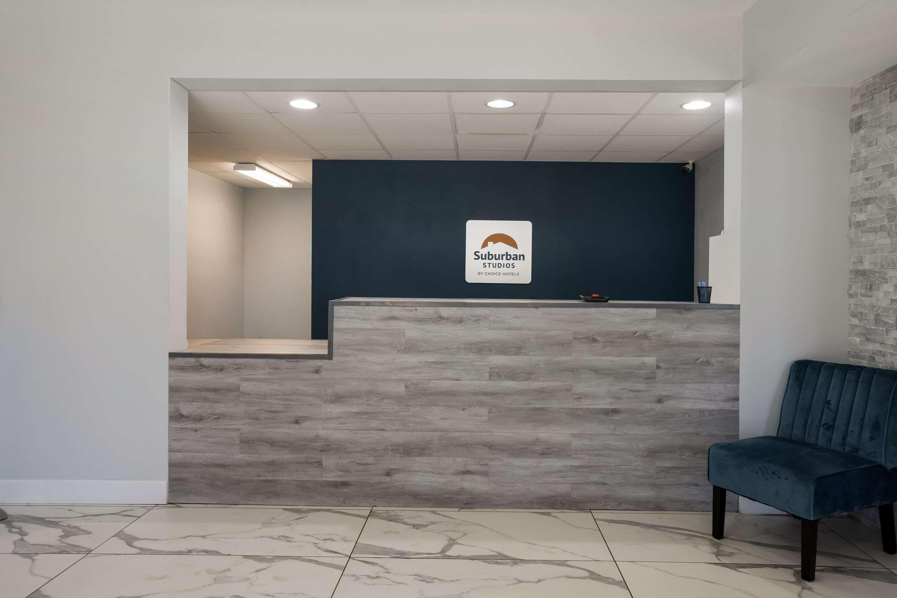 A reception desk with a sign for Suburban Studios by Choice Hotels, a waiting area with a blue chair, and a tiled floor.
