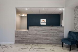 A reception desk with a sign for Suburban Studios by Choice Hotels, a waiting area with a blue chair, and a tiled floor. - Suburban Studios Hinesville - Fort Stewart Area (Hinesville)