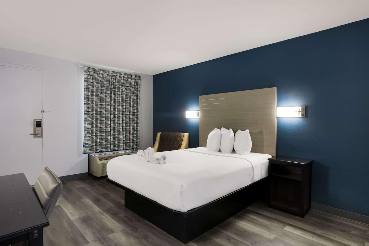 Una habitación de hotel con cama, mesitas de noche, escritorio, silla, ventana con cortinas e iluminación en la pared.