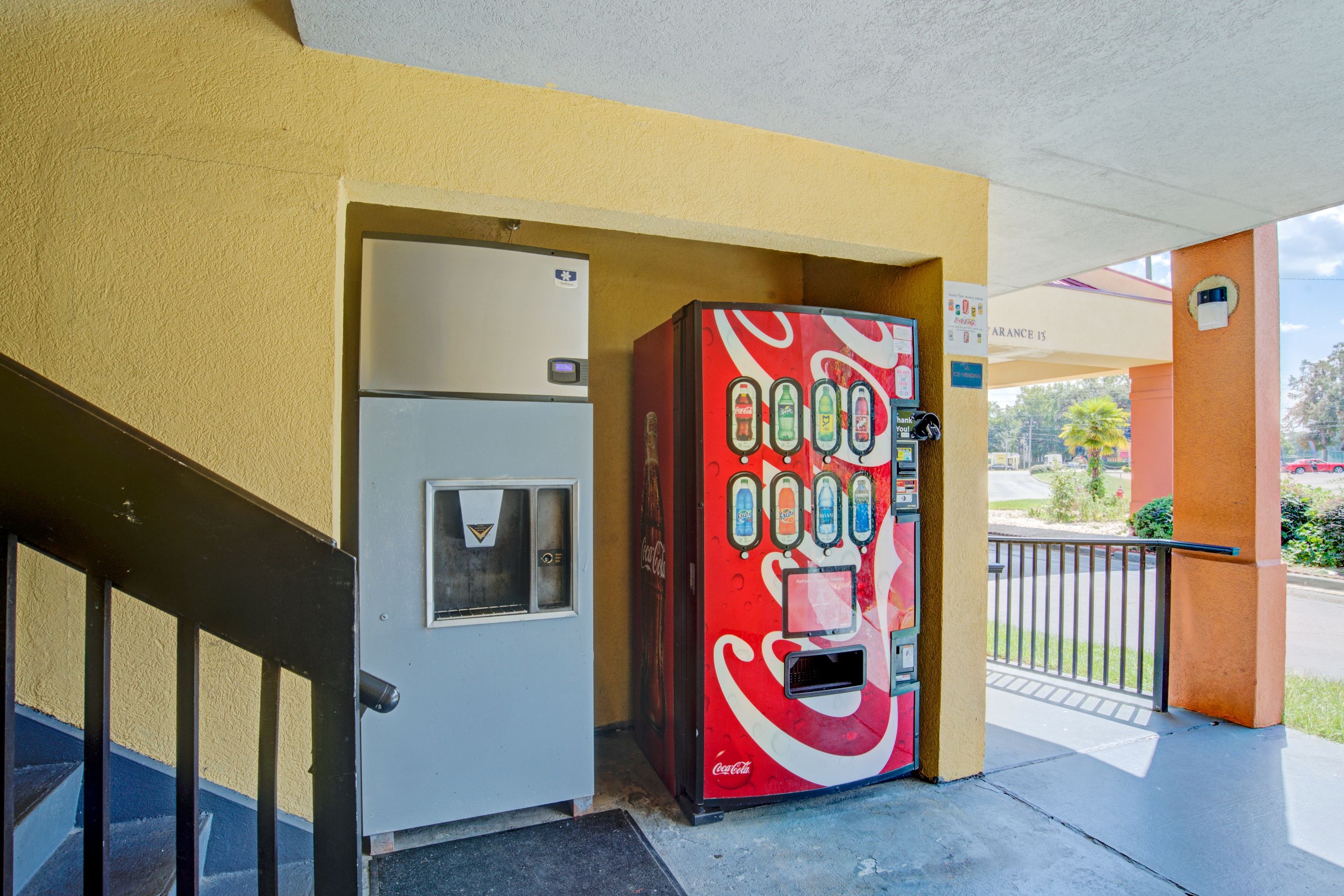 Una máquina expendedora de Coca-Cola junto a una puerta, con una escalera y una zona cubierta al fondo.