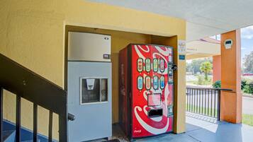 Una máquina expendedora de Coca-Cola junto a una puerta, con una escalera y una zona cubierta al fondo.