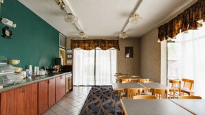 A dining area with tables and chairs, a counter with food containers, and a patterned carpet. - Suburban Studios Hinesville - Fort Stewart Area (Hinesville)