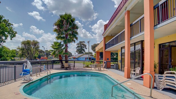 A swimming pool with a lifeguard chair, a palm tree, and a building with balconies and a storefront. - Suburban Studios Hinesville - Fort Stewart Area (Hinesville)