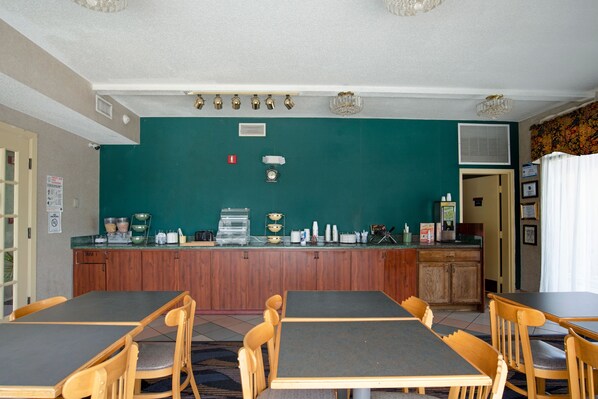 A dining area with wooden tables and chairs, a bar counter with various items, and a green wall. - Suburban Studios Hinesville - Fort Stewart Area (Hinesville)