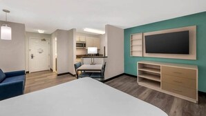 Une chambre d’hôtel moderne avec un grand lit, un bureau, une chaise, une télévision et une armoire. On aperçoit également une kitchenette au fond de la pièce.