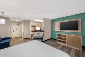 Una habitación de hotel moderna con una cama grande, un escritorio, una silla, un televisor, un armario y una kitchenette al fondo.