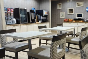 A hotel dining area with white tables and patterned chairs, a refrigerator, coffee machines, and a fruit display. - Sonesta Essential Hotels Overland Park (Overland Park)