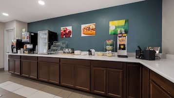 A hotel reception area with a counter, coffee machines, and a wall with fruit and breakfast items.