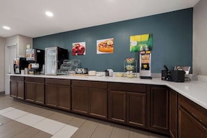 A hotel reception area with a counter, coffee machines, and a wall with fruit and breakfast items. - Quality Inn University Area Farmville, VA (Farmville)