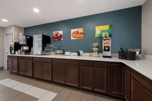 Zona de recepción de hotel con mostrador, máquinas de café y una pared decorada con frutas y productos para el desayuno.
