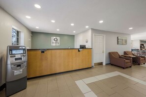 A hotel reception area with an ATM, a reception desk, a sign indicating 'Quality by Choice Hotels', and a seating area with wicker chairs.