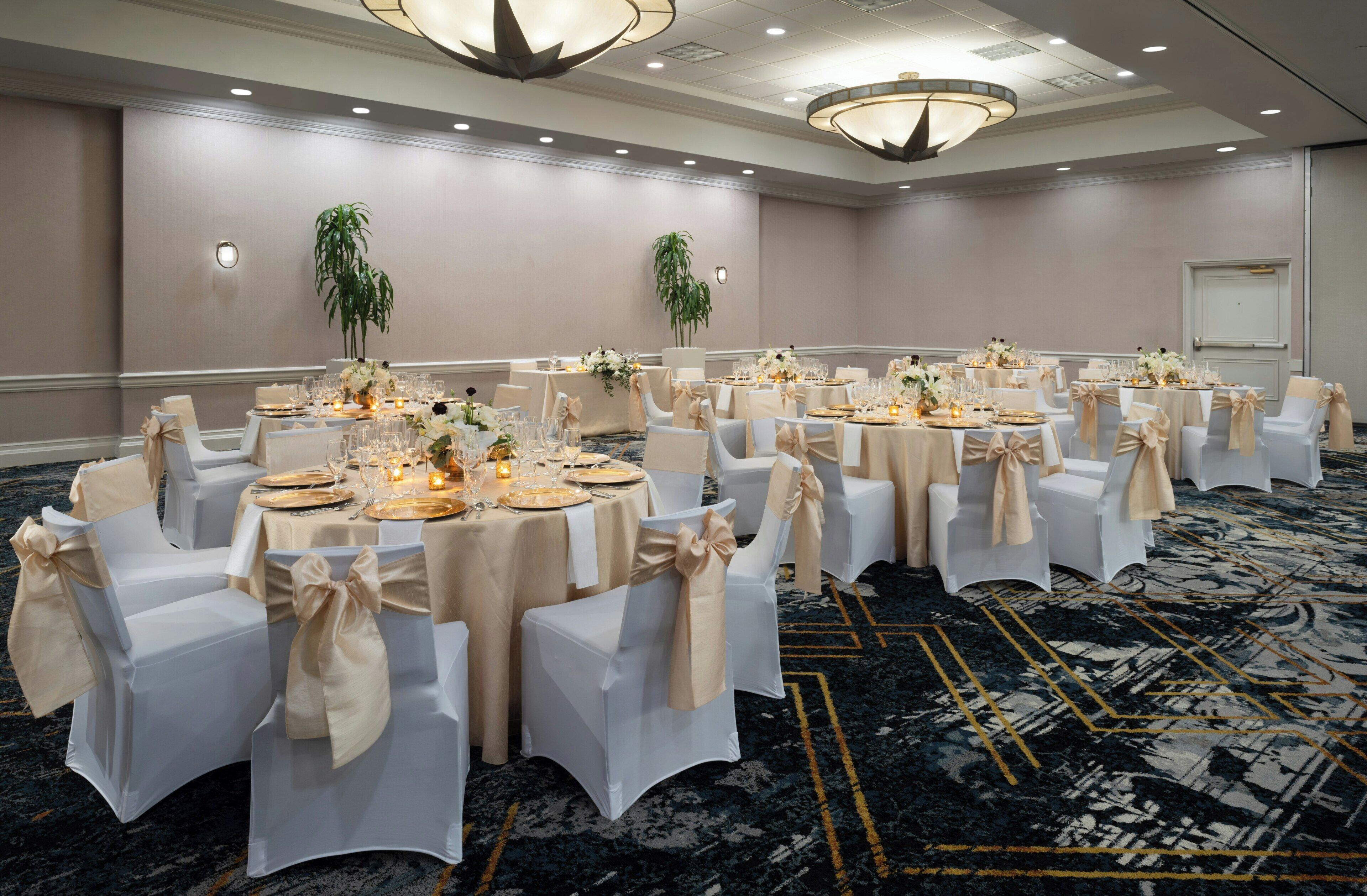A banquet hall with round tables, white chairs, and chandeliers.