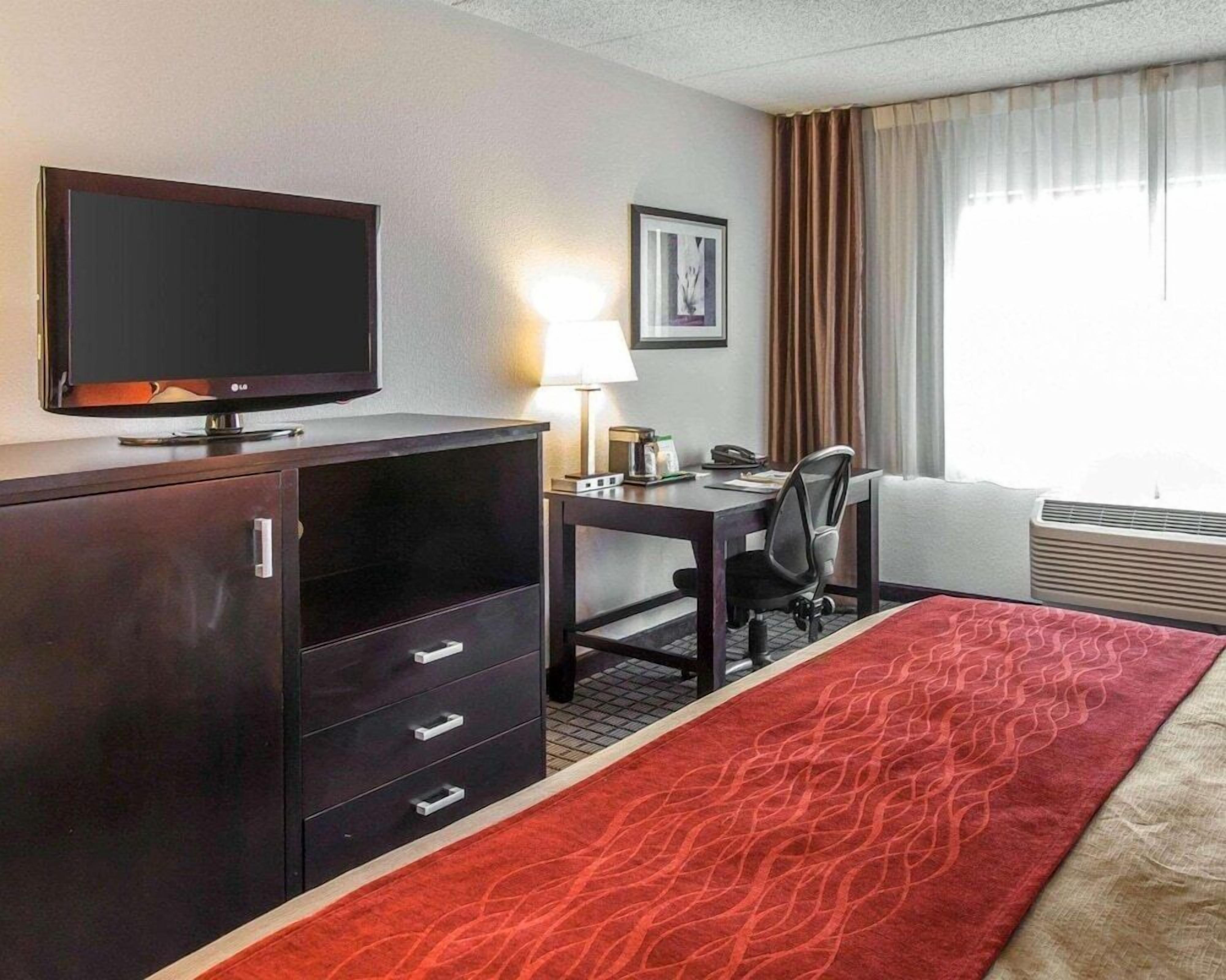 A hotel room with a TV, desk, chair, lamp, and a bed with a red patterned bedspread.