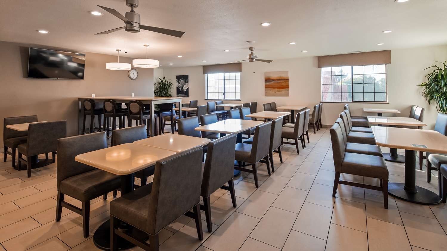 A spacious dining area with multiple tables and chairs, ceiling fans, and large windows providing natural light.