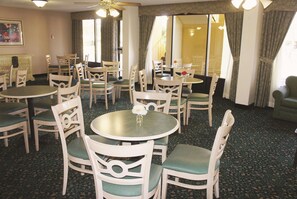 Comedor de hotel con varias mesas redondas y sillas, ventanas amplias con cortinas y un ventilador de techo.