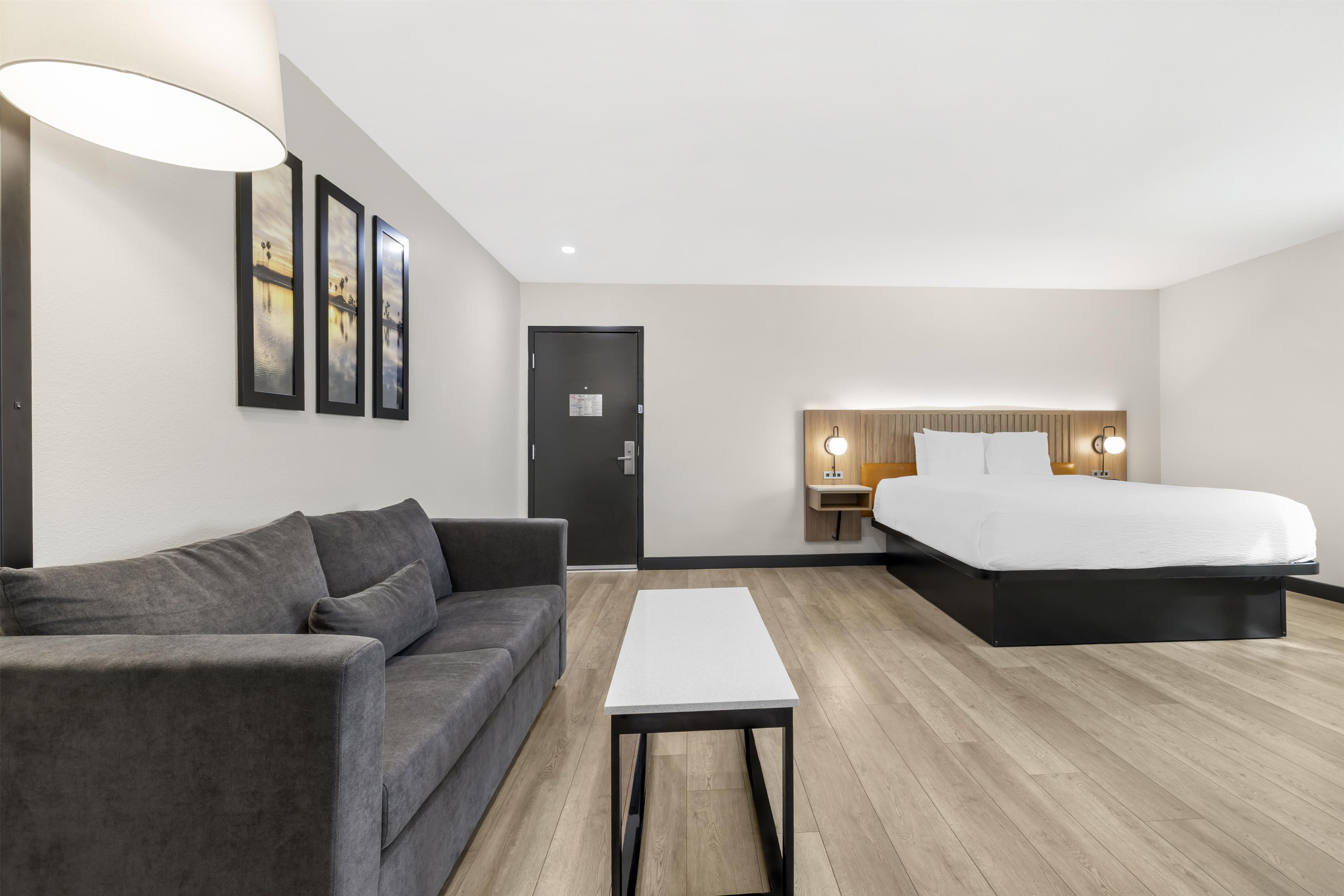 A modern hotel room with a grey sofa, a white table, a bed with a wooden headboard, and framed pictures on the wall.