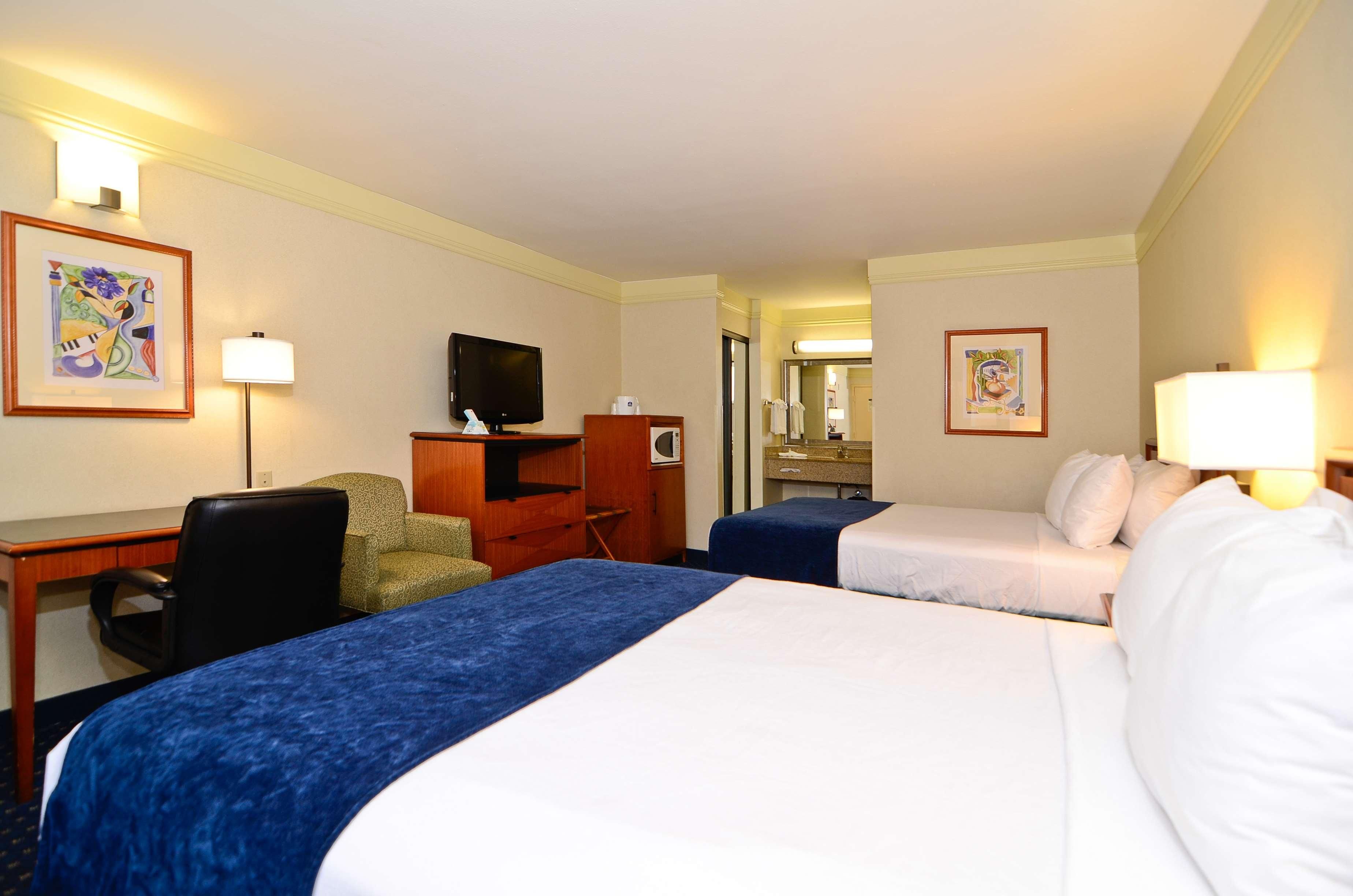 Habitación de hotel con dos camas, un escritorio con silla, televisor y cuadros en las paredes.