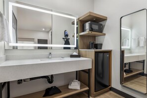 A modern bathroom with a white sink, a mirror, a black refrigerator, and a microwave on a wooden shelf.