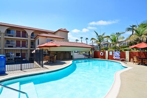 A swimming pool with a curved edge, a covered seating area with umbrellas, and a building with multiple balconies.