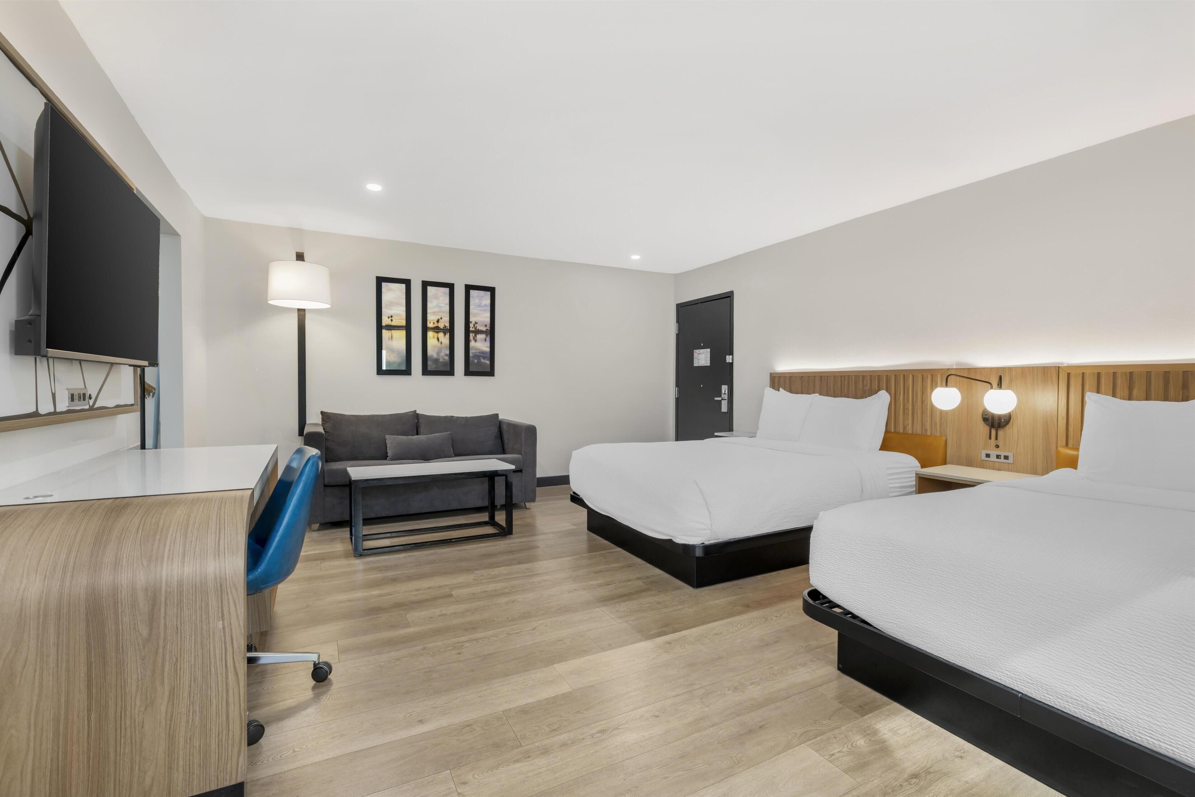 Ein Hotelzimmer mit Schreibtisch, zwei Betten, einer Couch, einem Fernseher und gerahmten Bildern an der Wand.