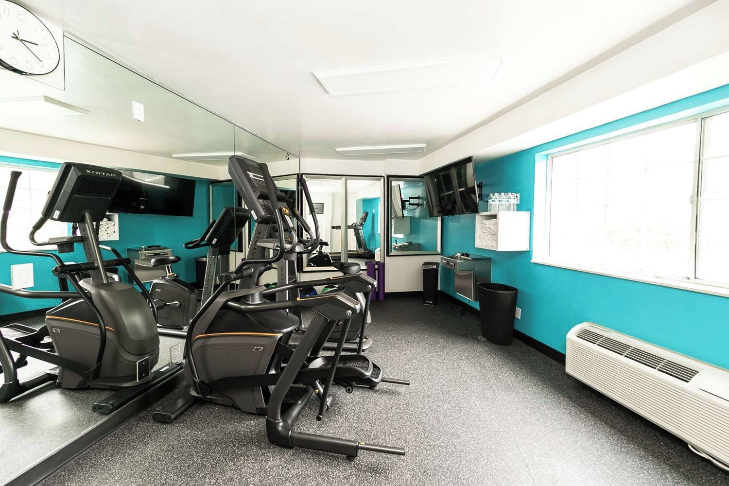 Une salle de sport équipée de matériel d’exercice, d’un miroir, d’une horloge et d’une fenêtre avec des stores.