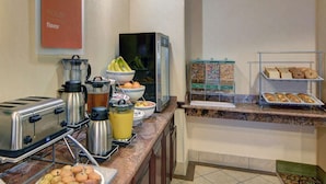 A breakfast bar with a coffee maker, toaster, and muffin tray. There are bowls of fruit and a refrigerator in the background. - Motel6 Fort Worth, TX I-35 & I-20 (Fort Worth)