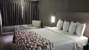 Une chambre d’hôtel avec un lit, une chaise, une table de chevet, une lampe et une fenêtre avec des rideaux.