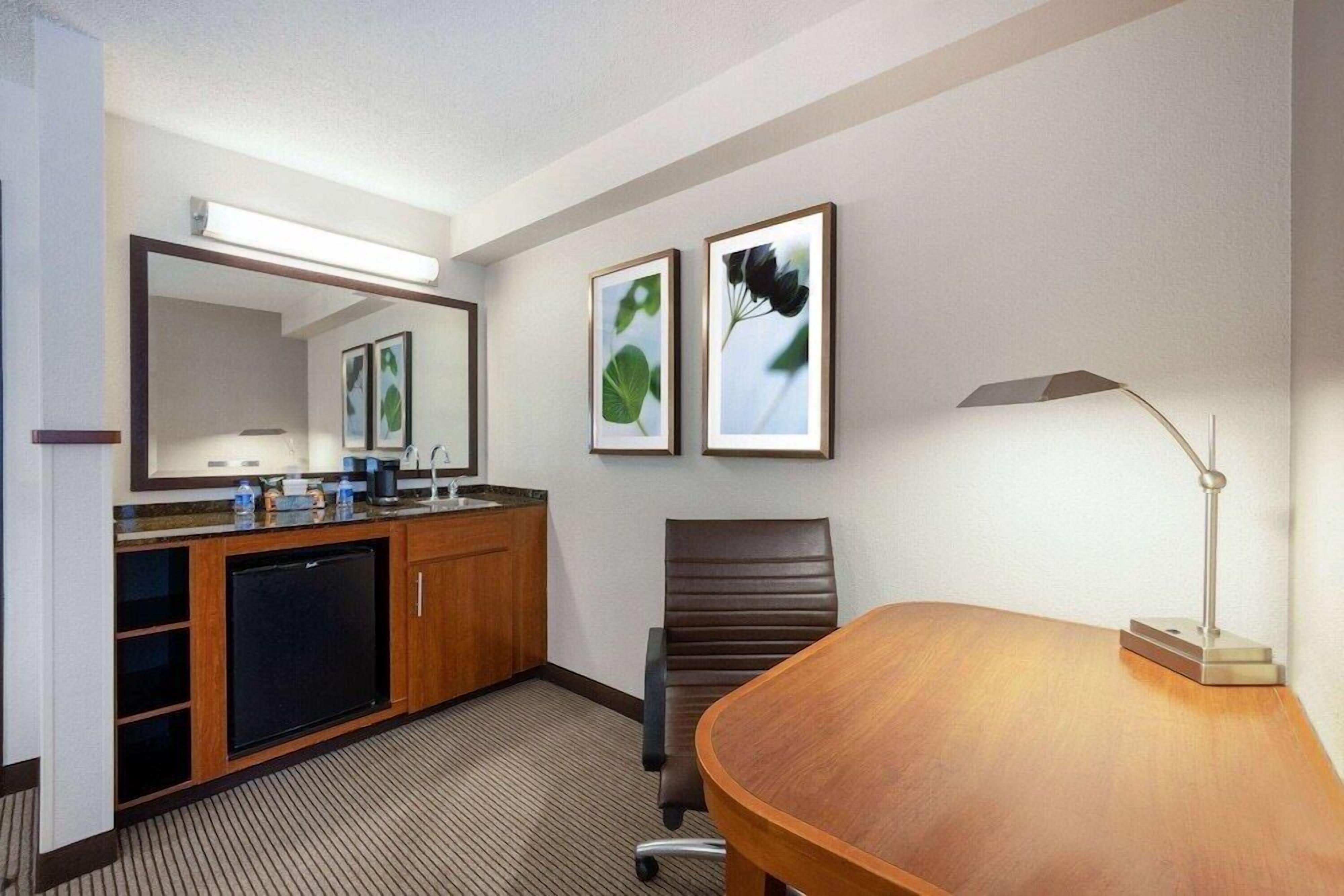 Habitación de hotel con un escritorio de madera, una silla, una lámpara de escritorio, un espejo, un lavamanos y cuadros en la pared.