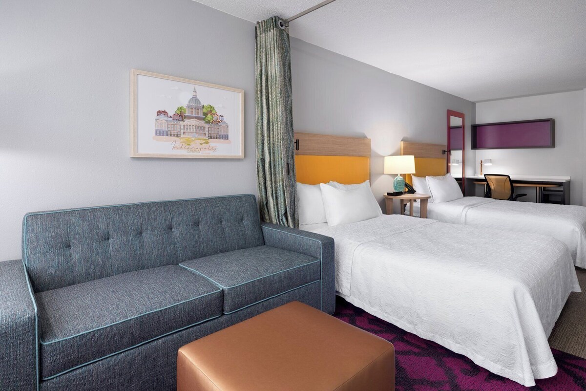 Habitación de hotel con un sofá gris, dos camas, un escritorio, una lámpara y un cuadro con una imagen de un edificio.