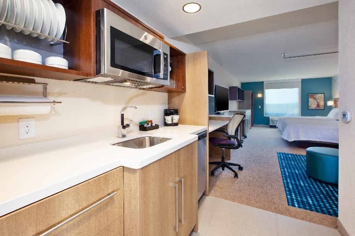 A compact hotel room with a kitchenette, a bed, a desk, and a view of the ocean.