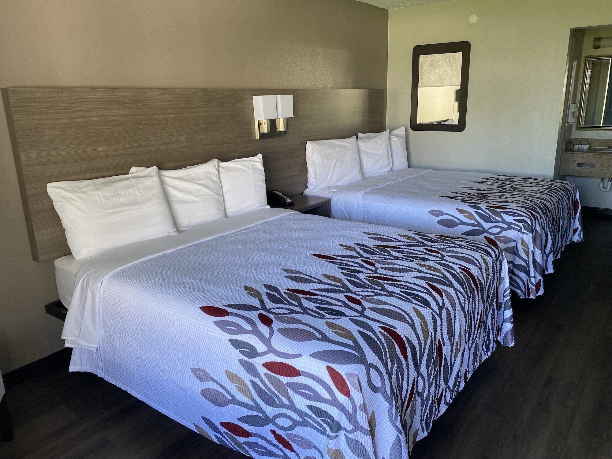 A hotel room with two beds, each with white linens and a patterned bedspread. There is a nightstand with a phone and a wall-mounted lamp.