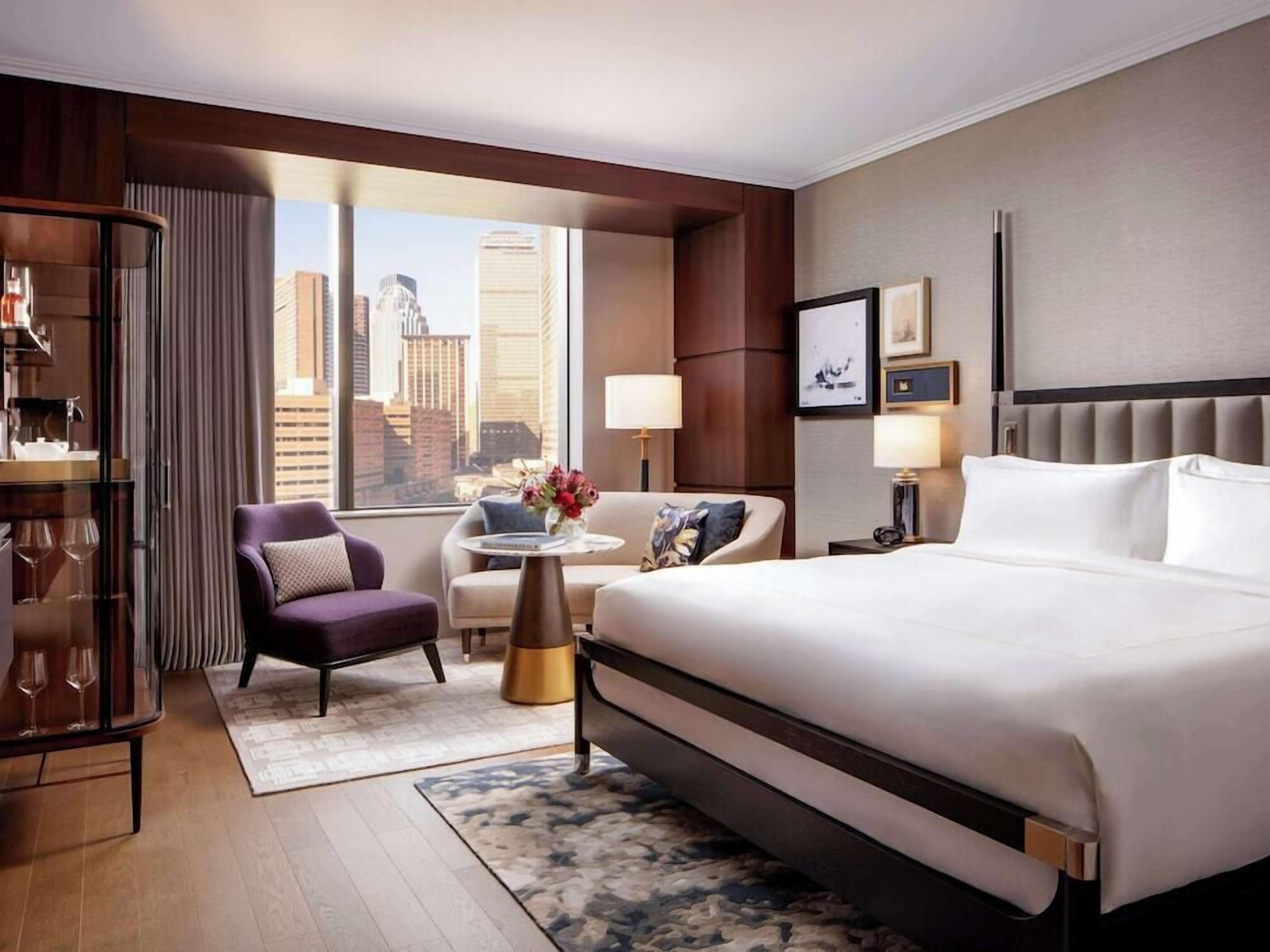 A modern hotel room with a large bed, a seating area with a small table, and a view of the city skyline.