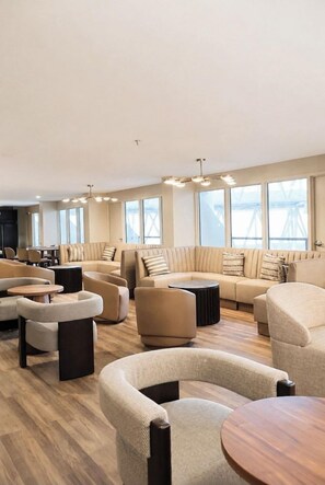 Ein moderner Loungebereich mit Sofas und Sesseln, die um einen Couchtisch angeordnet sind, große Fenster sorgen für natürliches Licht und bieten einen Blick ins Freie.