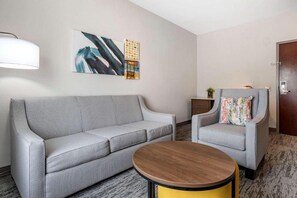 A hotel room with a grey sofa, a round wooden table, a grey armchair with a colorful pillow, and a wall with abstract art. - Tryp by Wyndham Tallahassee North I-10 Capital Cir (Tallahassee)