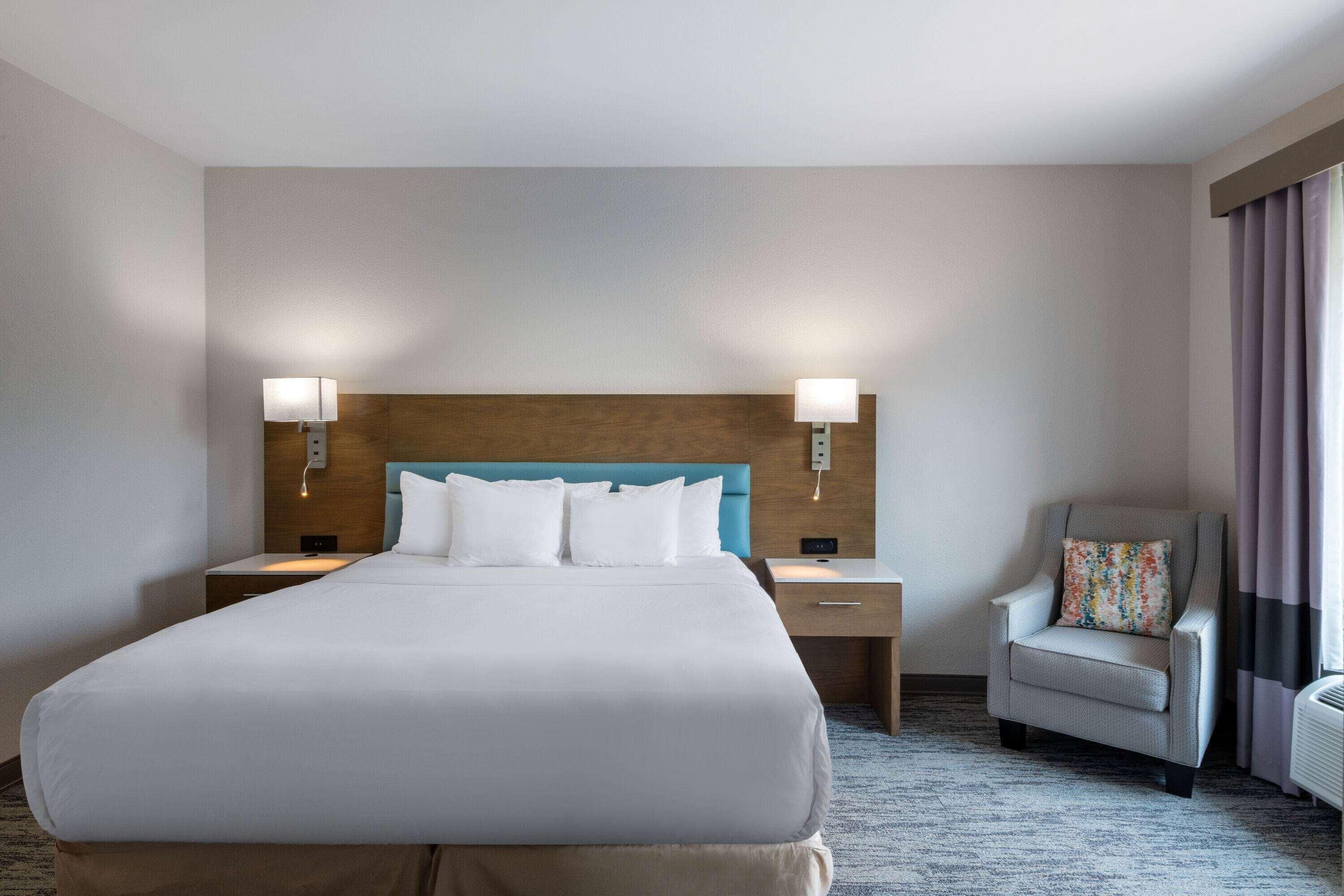 Habitación de hotel con una cama grande, dos mesitas de noche, una silla y un cabecero con lámparas de pared.