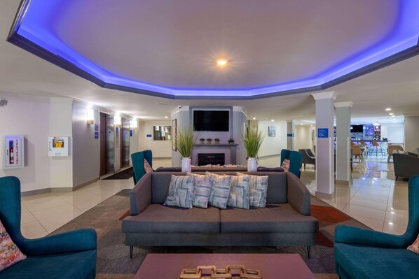 A modern lobby with a grey sofa, teal armchairs, a flat-screen TV, and a fireplace. - Tryp by Wyndham Tallahassee North I-10 Capital Cir (Tallahassee)